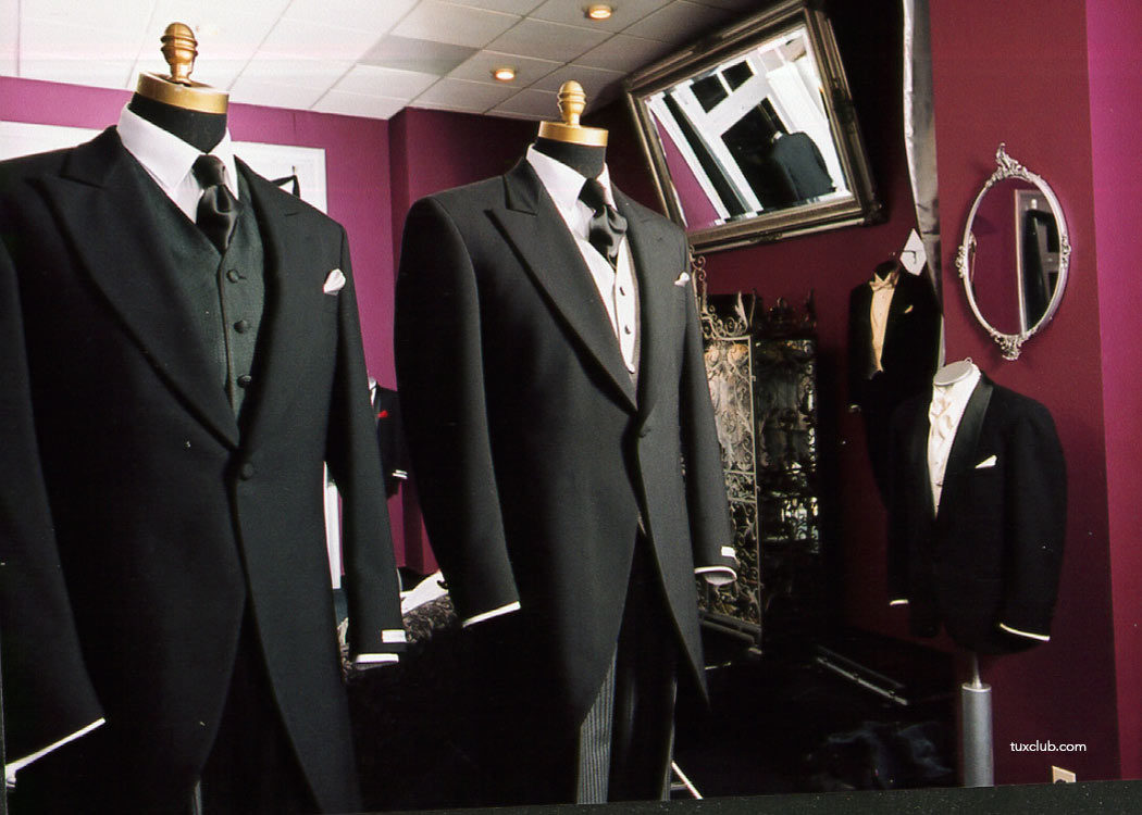 Two black cutaway morning suits on mannequins against a purple wall with a mirror inside a tuxedo shop