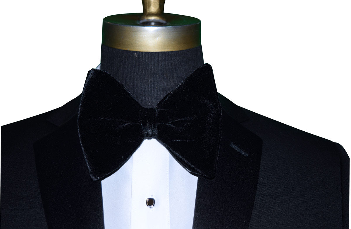 Black larege velvet bow tie on a mannequin wearing a black tuxedo against a white background
