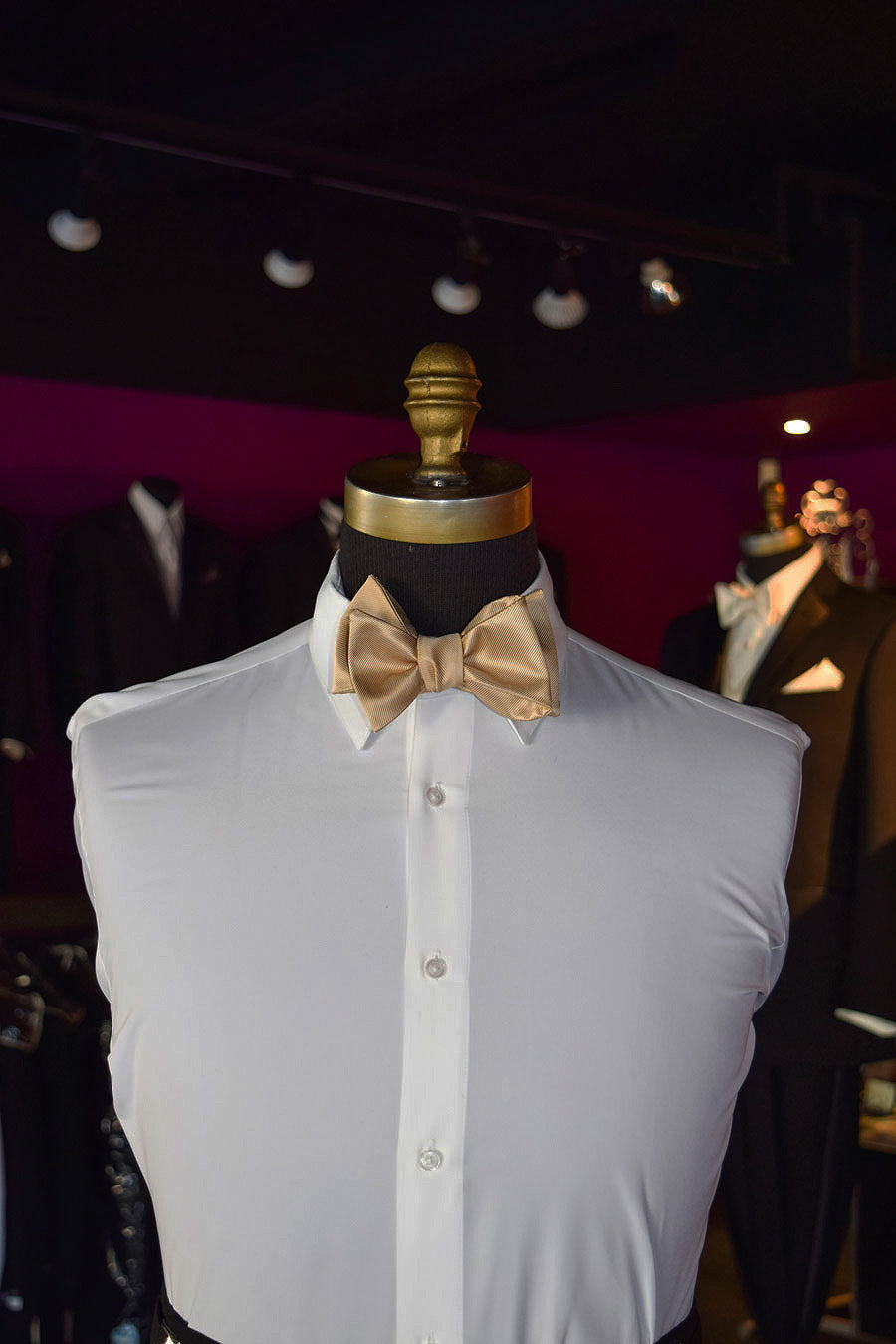 Mannequin wearing a white shirt with a gold "tie-yourself" bow tie inside of a tuxedo shop with mannequins in the background
