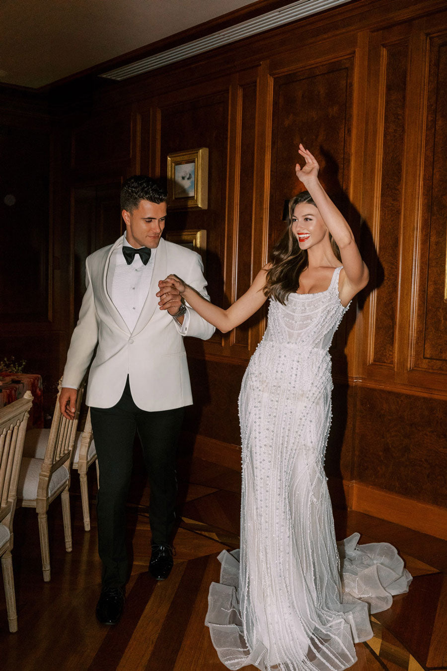 Groom in an off-white shawl collar tuxedo   and a bride in a off-white dress dancing in a dining room with wooden paneling.