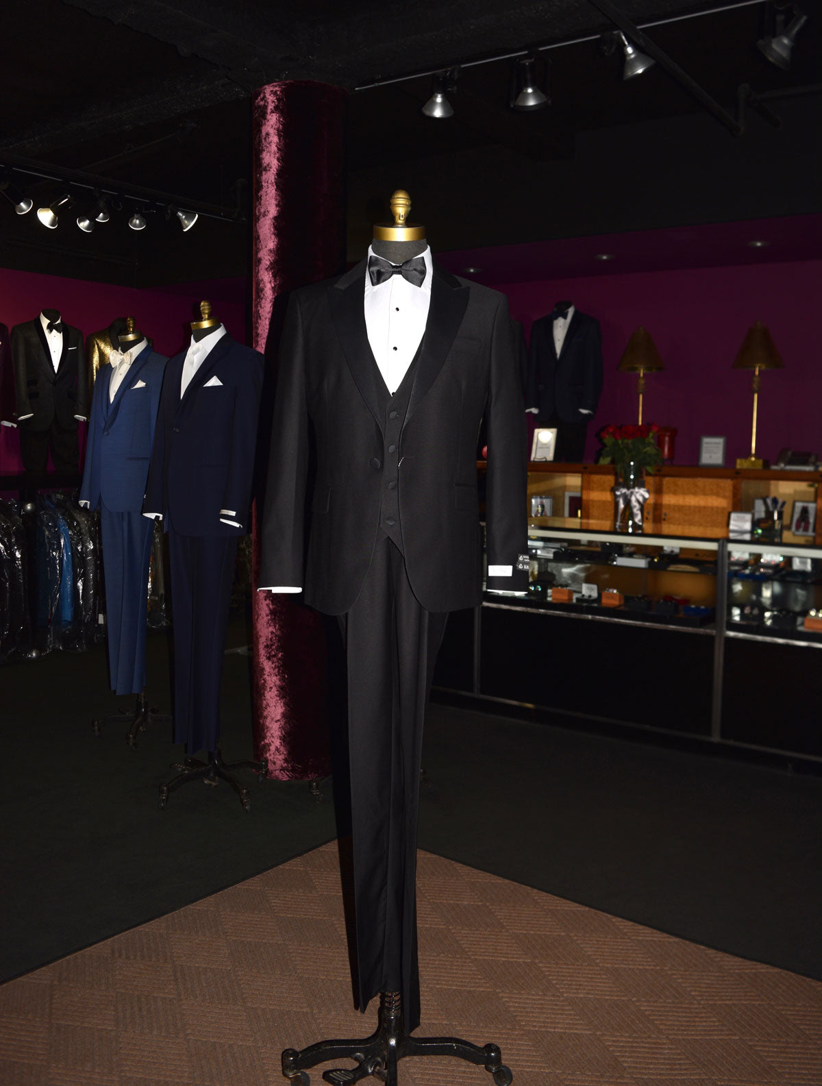 Black 3-piece slim-fit peak lapel tuxedo on a mannequin in a store setting with other suits and decor.