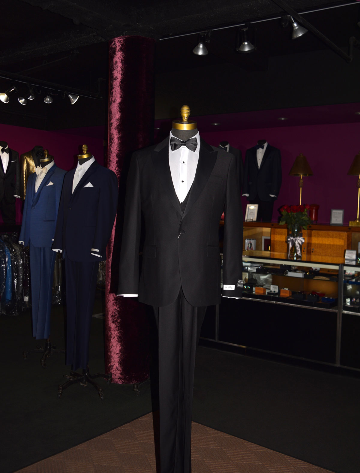 Black slim-fit 3 piece tuxedo on a mannequin in a formal wear store with other suits in the background.