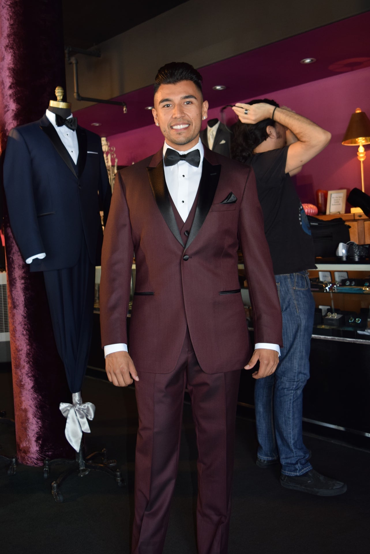 Man wearing a burgundy tuxedo and a burgundy vest with a black bow tie in a room with suits on display.