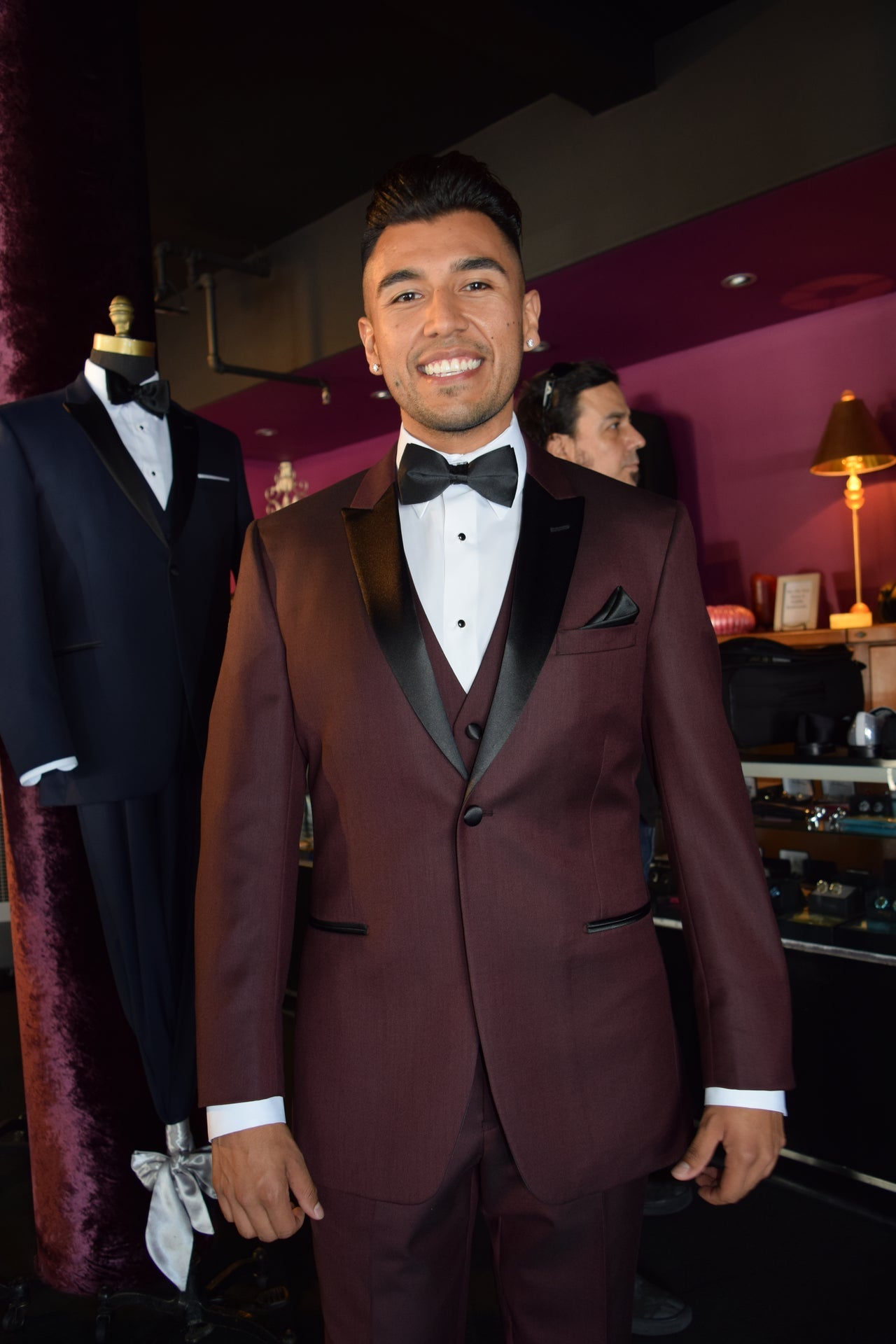 Man wearing a burgundy tuxedo with a burgundy vest in a fancy tuxedo shop with a mannequins on display.