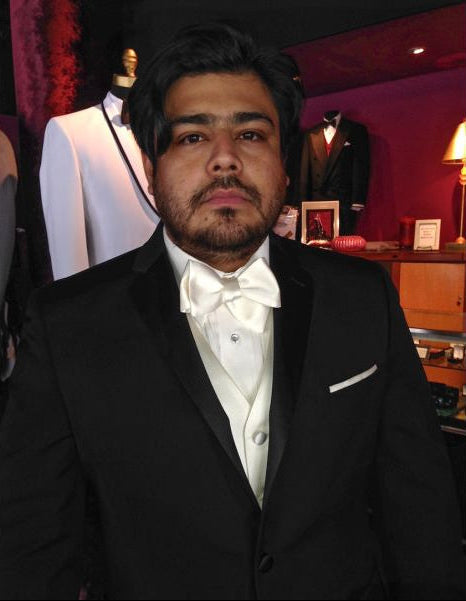 groom wearing a black tuxedo with an ivory "tie-yourself" bowtie inside a tuxedo shop ith tuxedos in the background