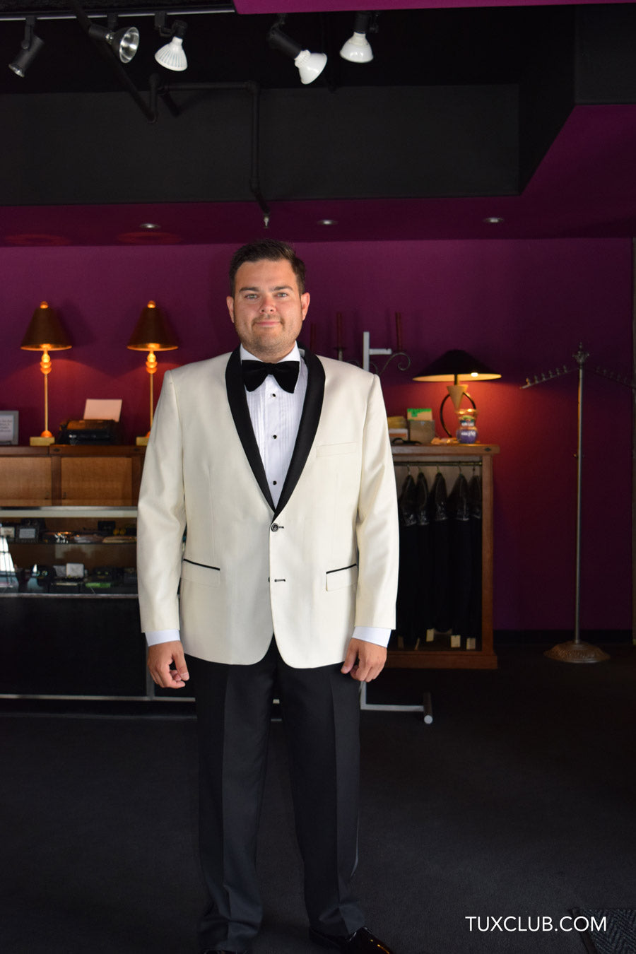 Man wearing an ivory tuxedo with black shawl lapel with a black velvet bowtie and black tuxedo pants standing in a tuxedo shop