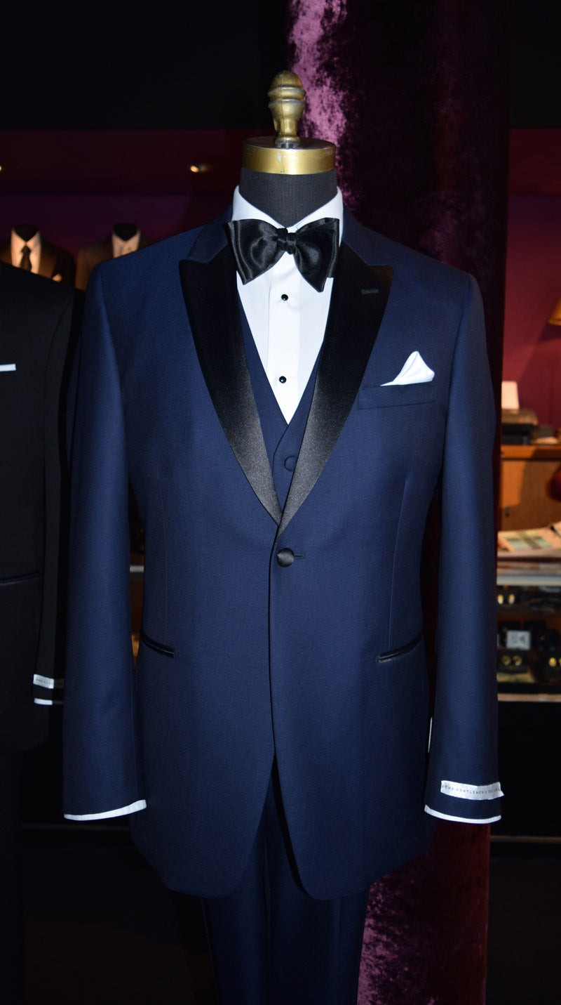 Navy blue tuxedo on a mannequin against with a navy blue vest and black bowtie on a mannequin inside a tuxedo shp