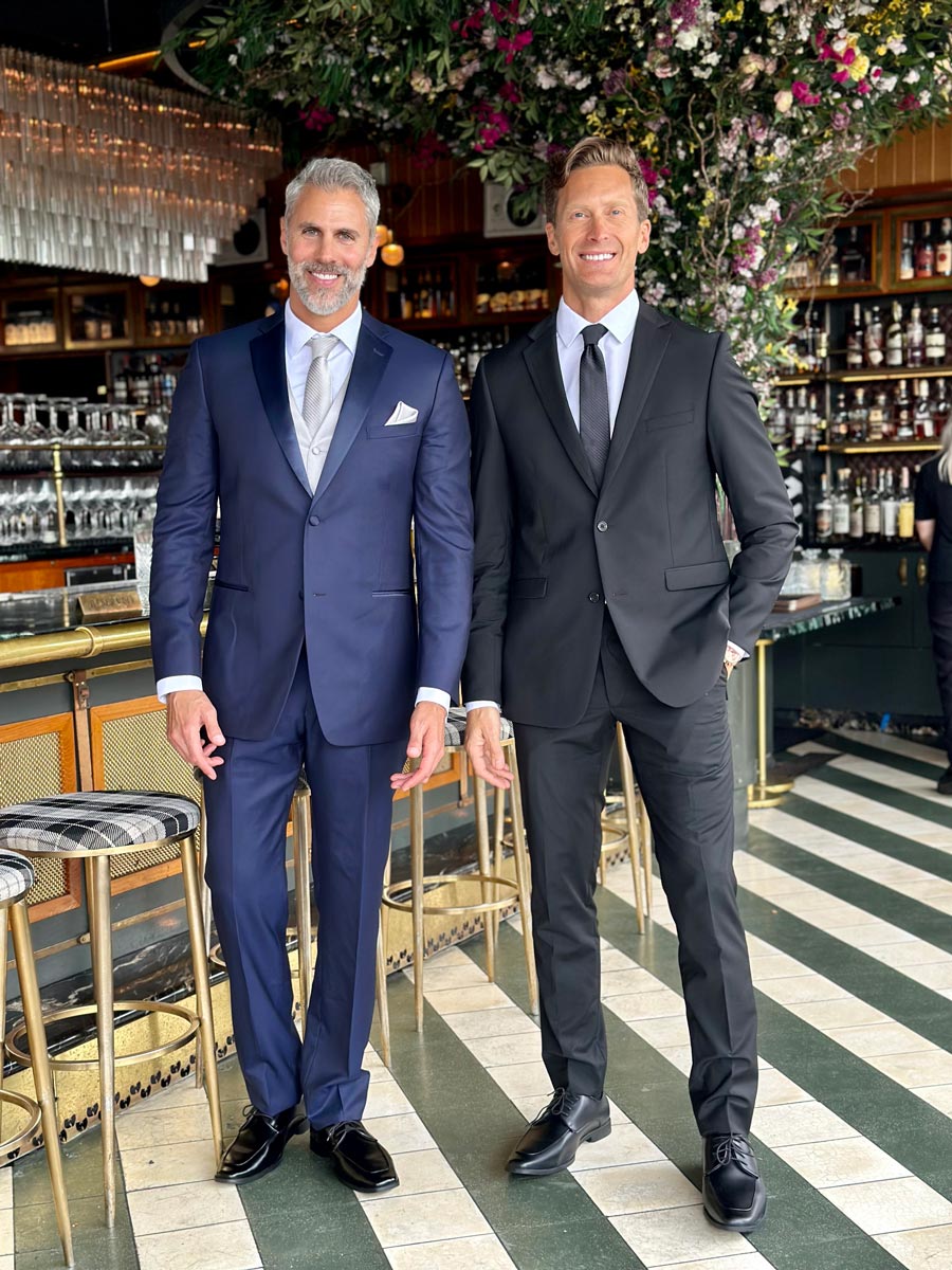 Two handsome men in suits at a hi-end restaurant. One is wearing a black slim fit suit and the other is wearing a navy blue tuxedo with blue lapels.