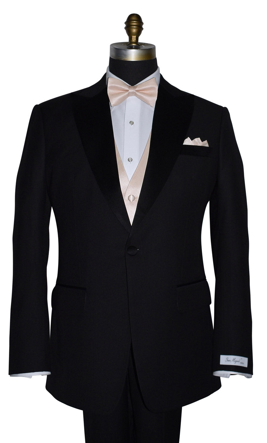 Black peak-lapel tuxedo with a "San Miguel Especial" branded label wearing a white shirt with a nude tuxedo vest and a nude bow tie on a mannequin against a white background