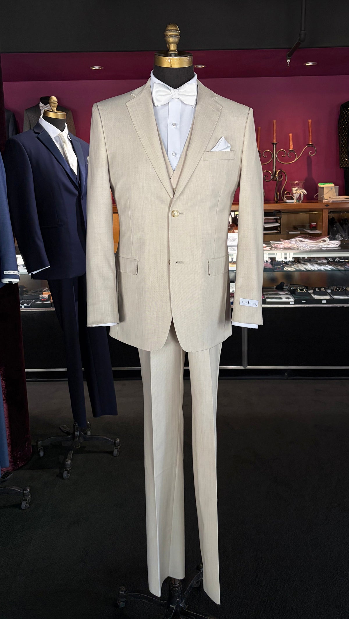 A tan fitted two-piece suit displayed on a mannequin torso, styled with a white dress shirt, white bow tie, and white pocket square, set against a dark boutique interior.