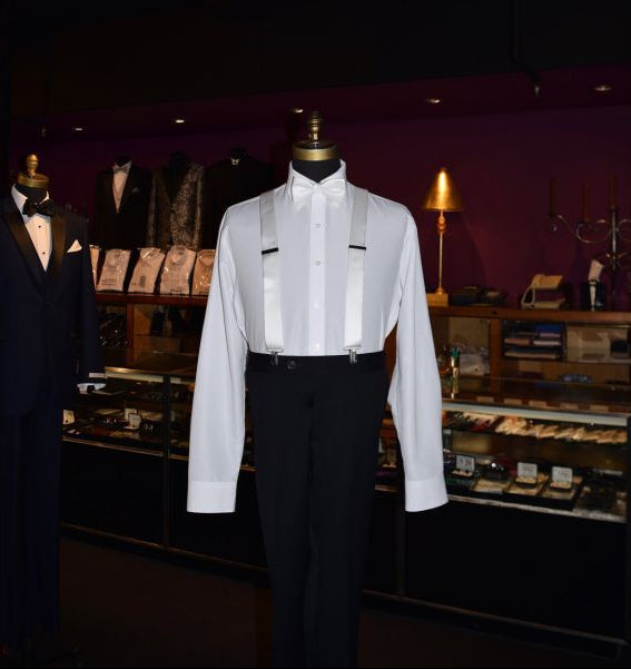 mannequin wearing a white tuxedo shirt, white suspenders and white bowtie on display inside a tuxedo shop with other tuxedos on display in the background 