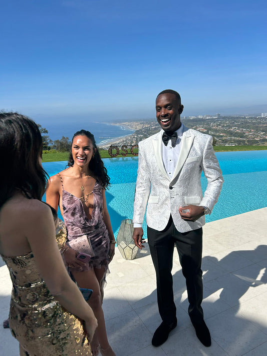 handsome man in a white peak lapel tuxedo with silver highlights and a black bowtie standing next to a pool with a scenic background